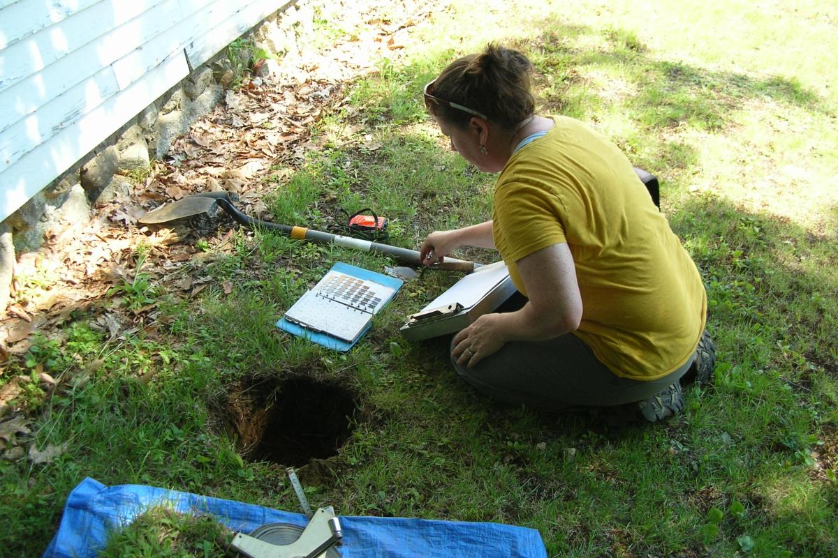 Archeologist Jessica Cofelice compares soil from the test pit to a soil color chart to determine its composition, and then records her observations.
